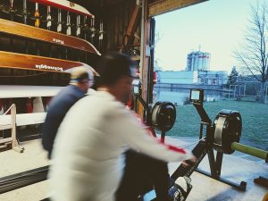 Zwei Personen trainieren auf Rudergeräten in einem Bootshaus. Durch die offene Tür ist eine ruhige Außenlandschaft sichtbar. Die Kombination aus körperlicher Anstrengung und der friedlichen Umgebung vermittelt eine interessante Atmosphäre.