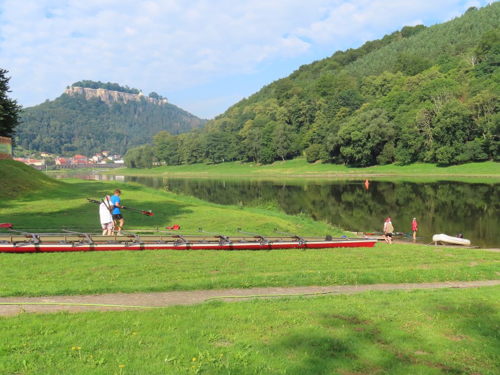 Eine sehr grüne Wiese an einem Fluss, darauf ein Ruderboot und einige Personen. Am gegenüberliegenden Ufer befindet sich ein bewaldeter Hügel, im Hintergrund eine Burg.