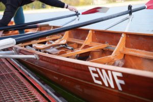 Das Bild zeigt ein Ruderboot aus Holz, das am Ufer eines Gewässers liegt. Es handelt sich um eine Nahaufnahme des Bootsinneren, wobei die Sitze und Ruderhalterungen sichtbar sind. Die Riemen sind schwarz mit rot-weißen Enden. Am Bug des Bootes sind die Buchstaben „EWF“ weiß aufgemalt. Eine Person ist teilweise zu sehen, wie sie die Ruder arrangiert. Die Umgebung scheint ein ruhiger Gewässerbereich mit grüner Vegetation im Hintergrund zu sein.